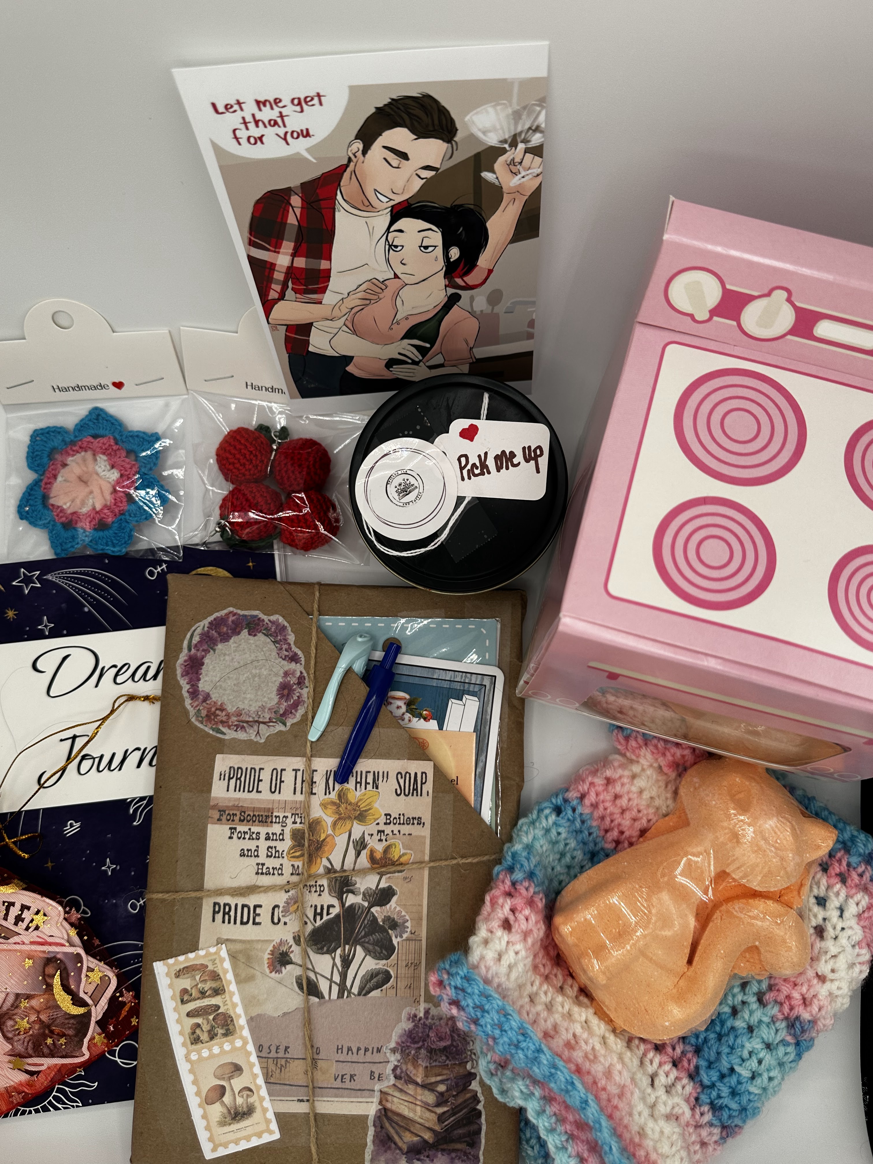 A flat lay of various items including a cartoon illustration card, crochet flowers, a black container with a label, a pink toy stove, a handcrafted journal with floral designs, a pen, and an orange cat-shaped bath bomb, arranged on a colorful knitted blanket.
